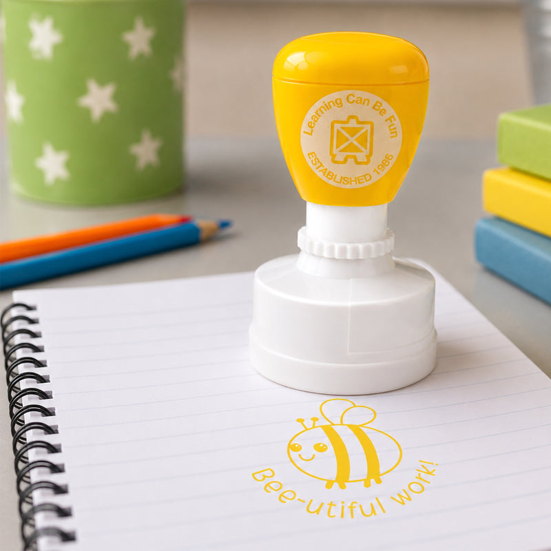 Learning Can Be Fun Bee-utiful Work self inking merit stamp positioned on a student notebook with a bright yellow bee themed impression. Classroom supplies, coloured pencils and books create an educational setting that highlights positive student recognition.