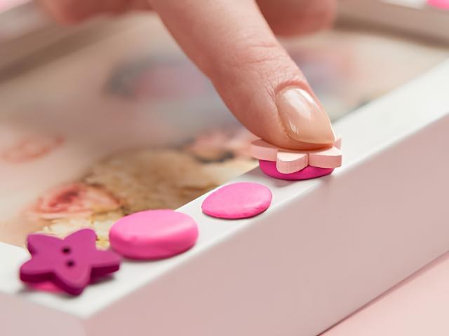 Fingertip pressing pink Blu Tack Colour shapes, including a star and a flower, along the edge of a white frame
