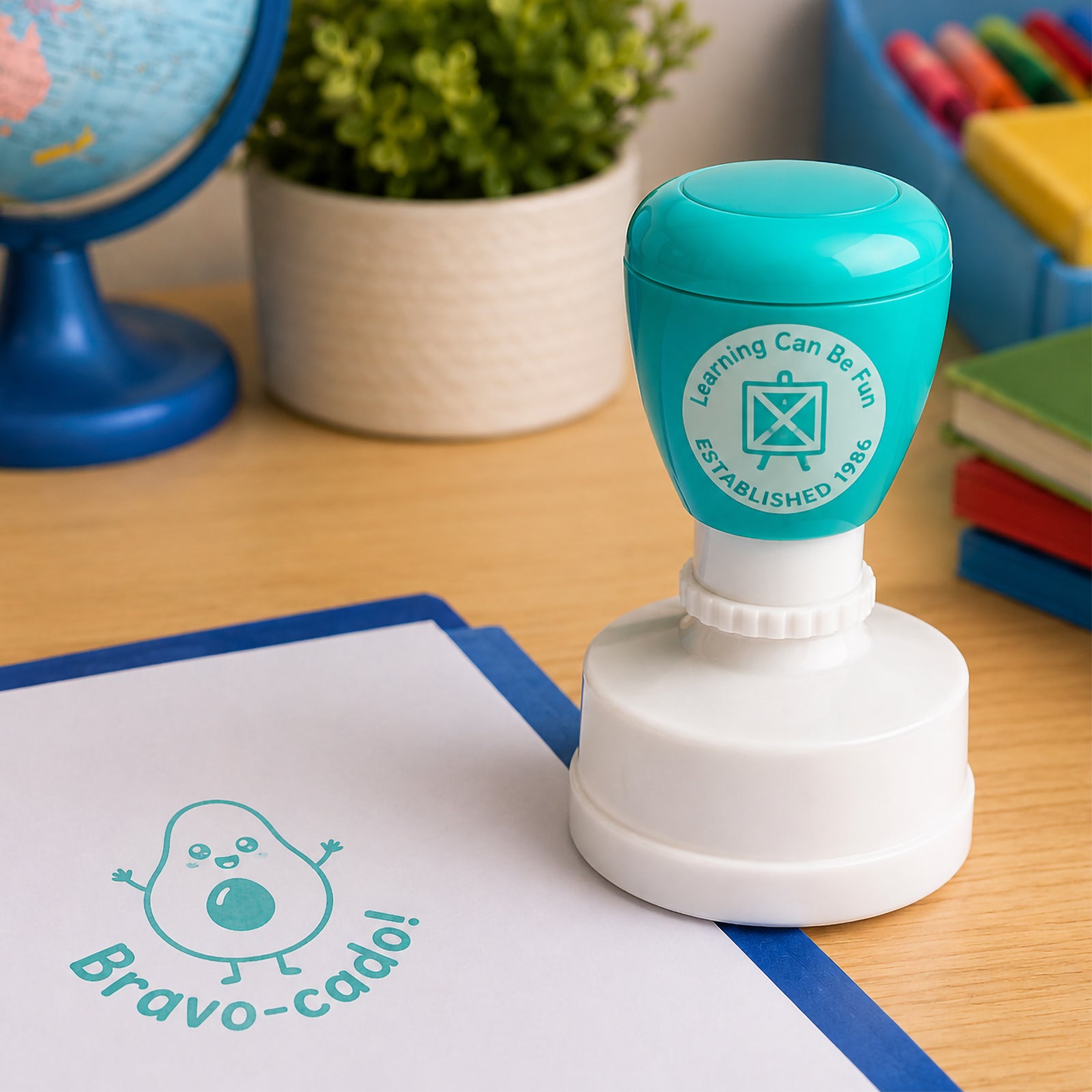 Close up classroom scene featuring the self inking merit stamp with a globe, colourful books and student materials. The cheerful avocado themed design helps celebrate effort, progress and learning milestones in an engaging way.