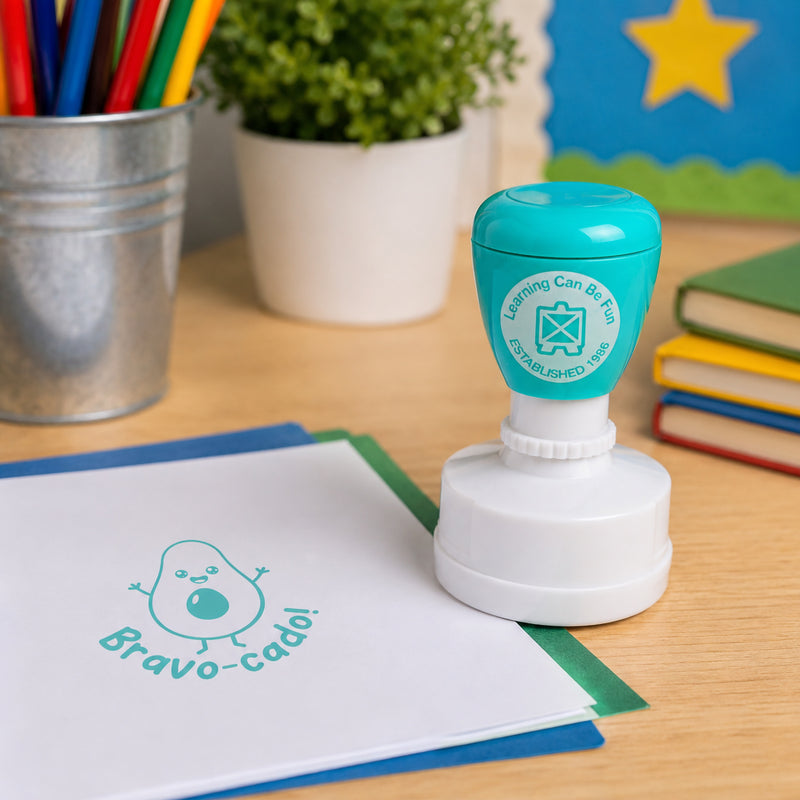 Aqua coloured Bravo-cado self inking merit stamp displayed on a classroom desk with books, coloured pencils and educational resources. The stamped impression shows a smiling avocado character and playful Bravo-cado! message, creating a fun way to recognise student achievement.