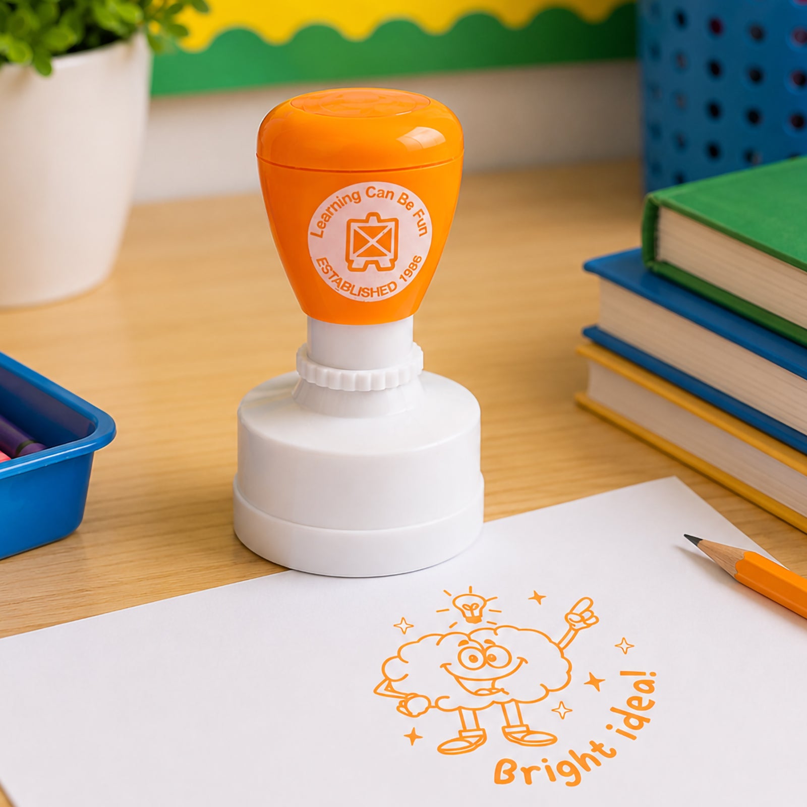 Classroom themed product image showing the self inking merit stamp beside books, pencils and educational supplies. The cheerful brain illustration and motivational message encourage confidence and creative learning.