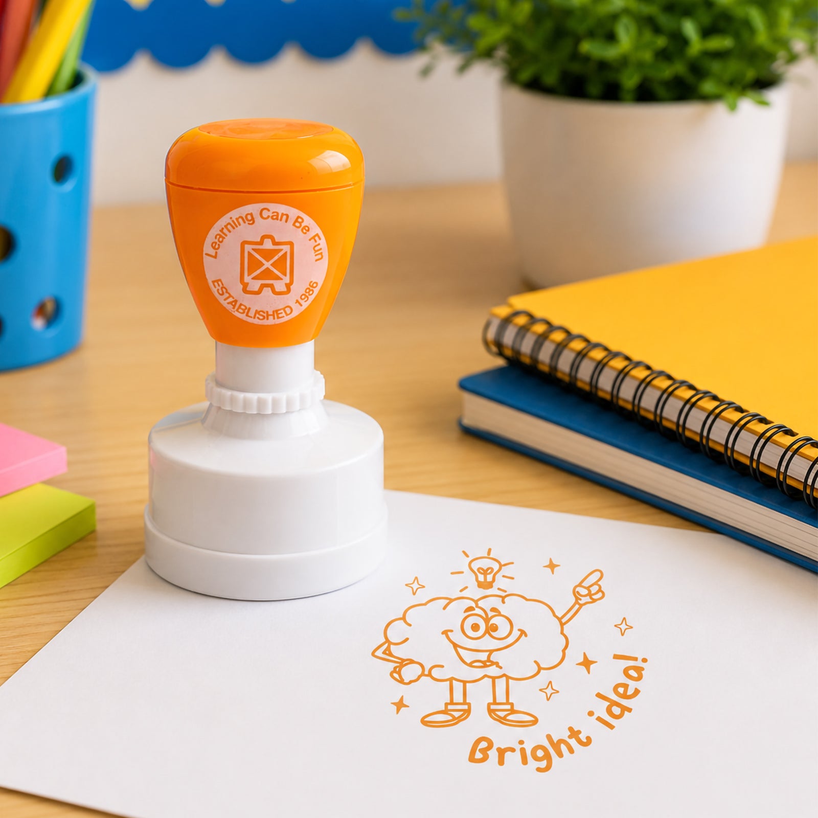 Orange Bright Idea reward stamp positioned on student work with colourful stationery and learning materials in the background. The bold impression highlights positive recognition for problem solving, participation and academic achievement.
