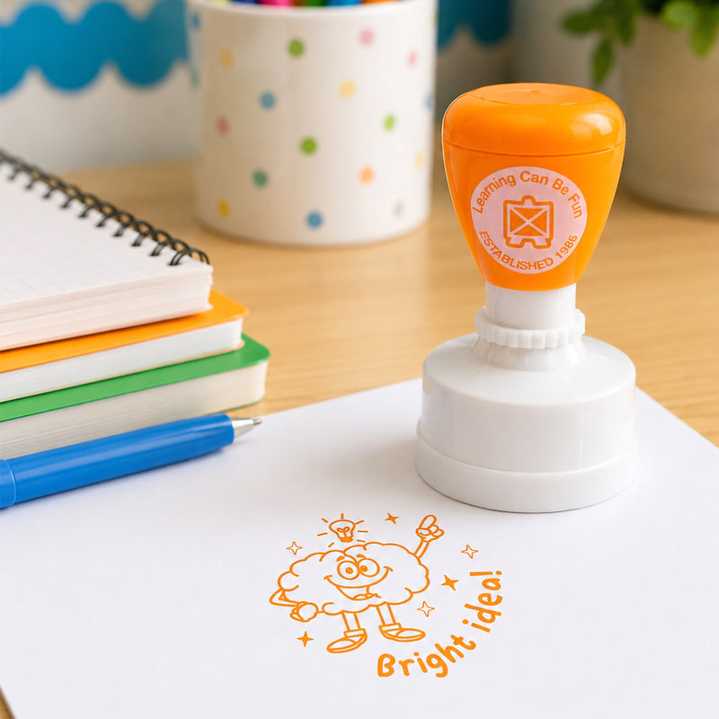 Learning Can Be Fun Bright Idea self inking merit stamp displayed on a classroom desk beside notebooks and stationery. The bright orange stamp impression features a smiling brain character with a lightbulb, celebrating creativity and innovative thinking.