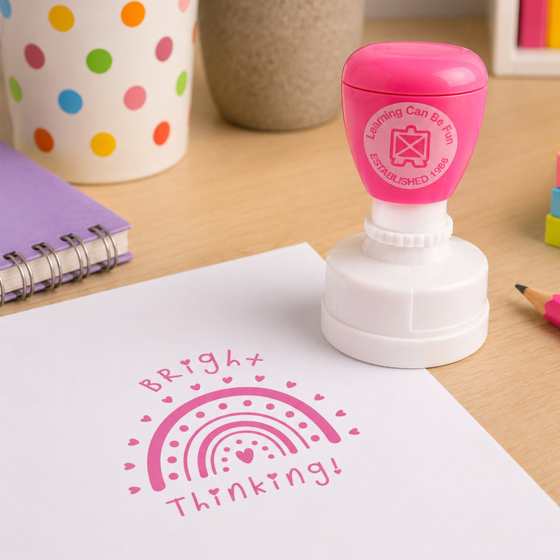 Learning Can Be Fun Bright Thinking self inking merit stamp displayed on a classroom desk beside notebooks and colourful stationery. The pink stamp impression features a decorative rainbow surrounded by hearts, celebrating creative thinking and student achievement.