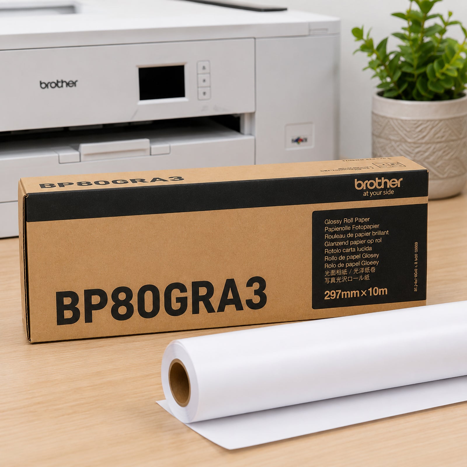 Brother BP80GRA3 glossy paper roll displayed beside genuine Brother packaging on an office desk with a compatible printer in the background. The premium 165gsm glossy paper roll is partially unrolled, highlighting its suitability for posters, promotional graphics and vibrant display printing.