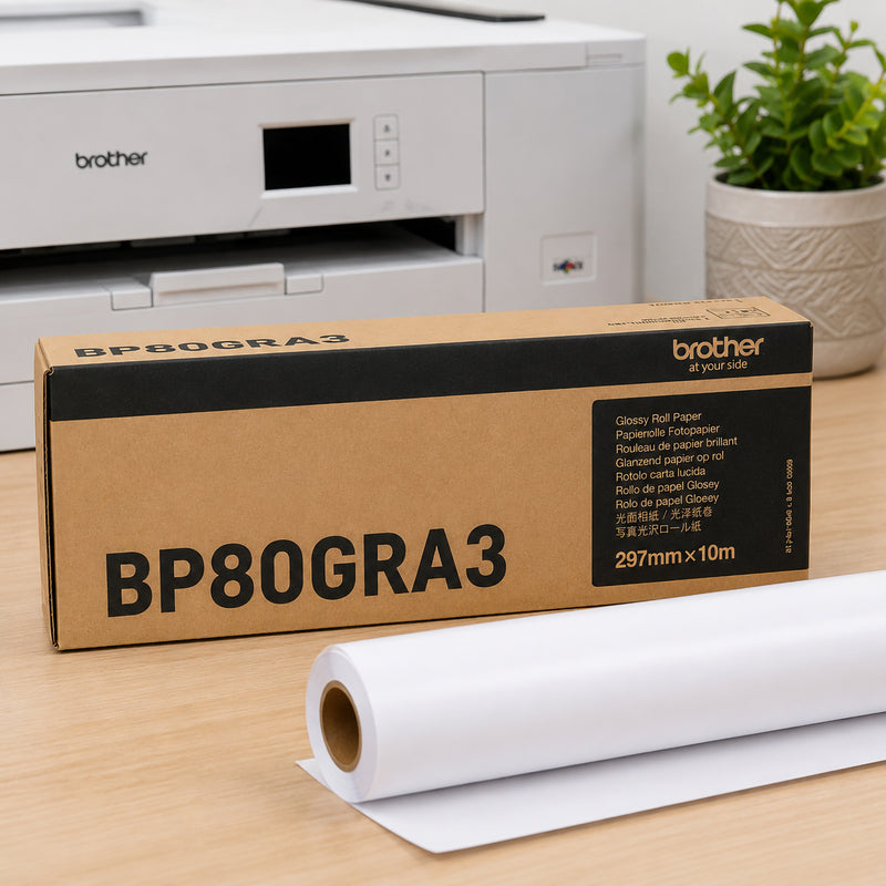 Brother BP80GRA3 glossy paper roll displayed beside genuine Brother packaging on an office desk with a compatible printer in the background. The premium 165gsm glossy paper roll is partially unrolled, highlighting its suitability for posters, promotional graphics and vibrant display printing.