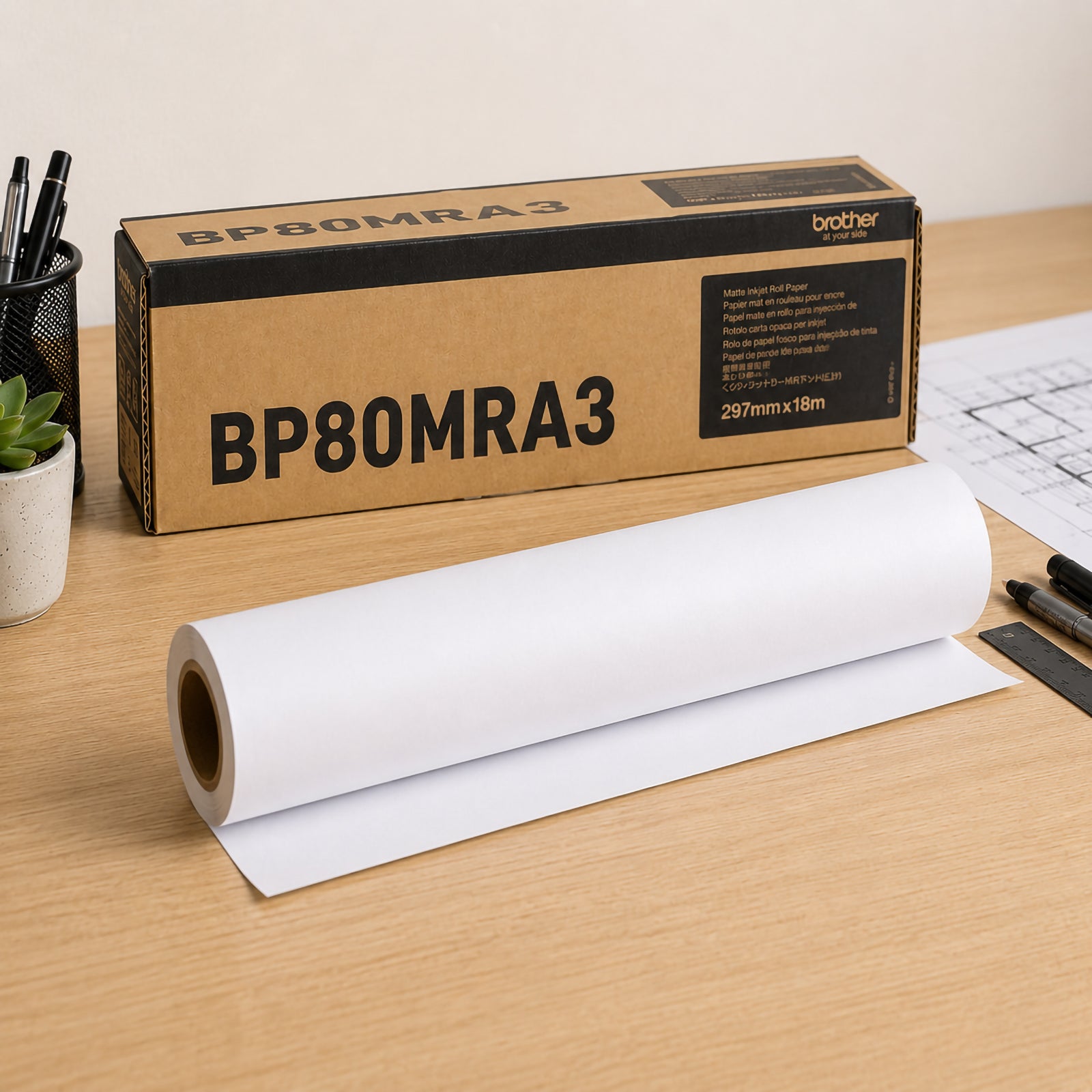 Brother BP80MRA3 matte paper roll displayed on a workstation desk beside its genuine Brother packaging. The premium white 145gsm paper roll is partially unrolled, with drafting tools and technical plans nearby, highlighting its suitability for professional signage, diagrams and presentation printing.