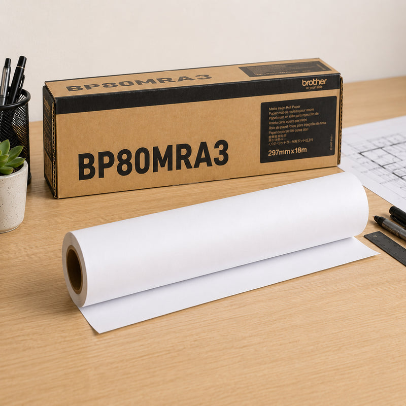 Brother BP80MRA3 matte paper roll displayed on a workstation desk beside its genuine Brother packaging. The premium white 145gsm paper roll is partially unrolled, with drafting tools and technical plans nearby, highlighting its suitability for professional signage, diagrams and presentation printing.