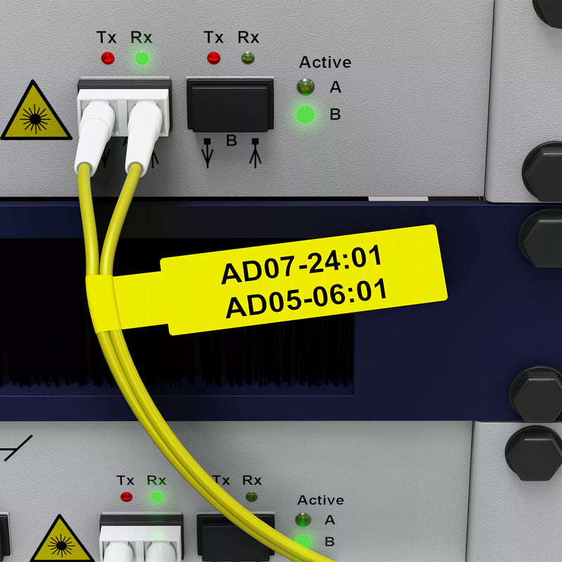 Brother FLe-6511 black on yellow flag labels attached around yellow fibre cables connected to professional network equipment. The bright yellow identification flag extends horizontally from the cables with bold black equipment references, demonstrating how the 21mm labels provide highly visible cable identification around densely configured technical hardware.