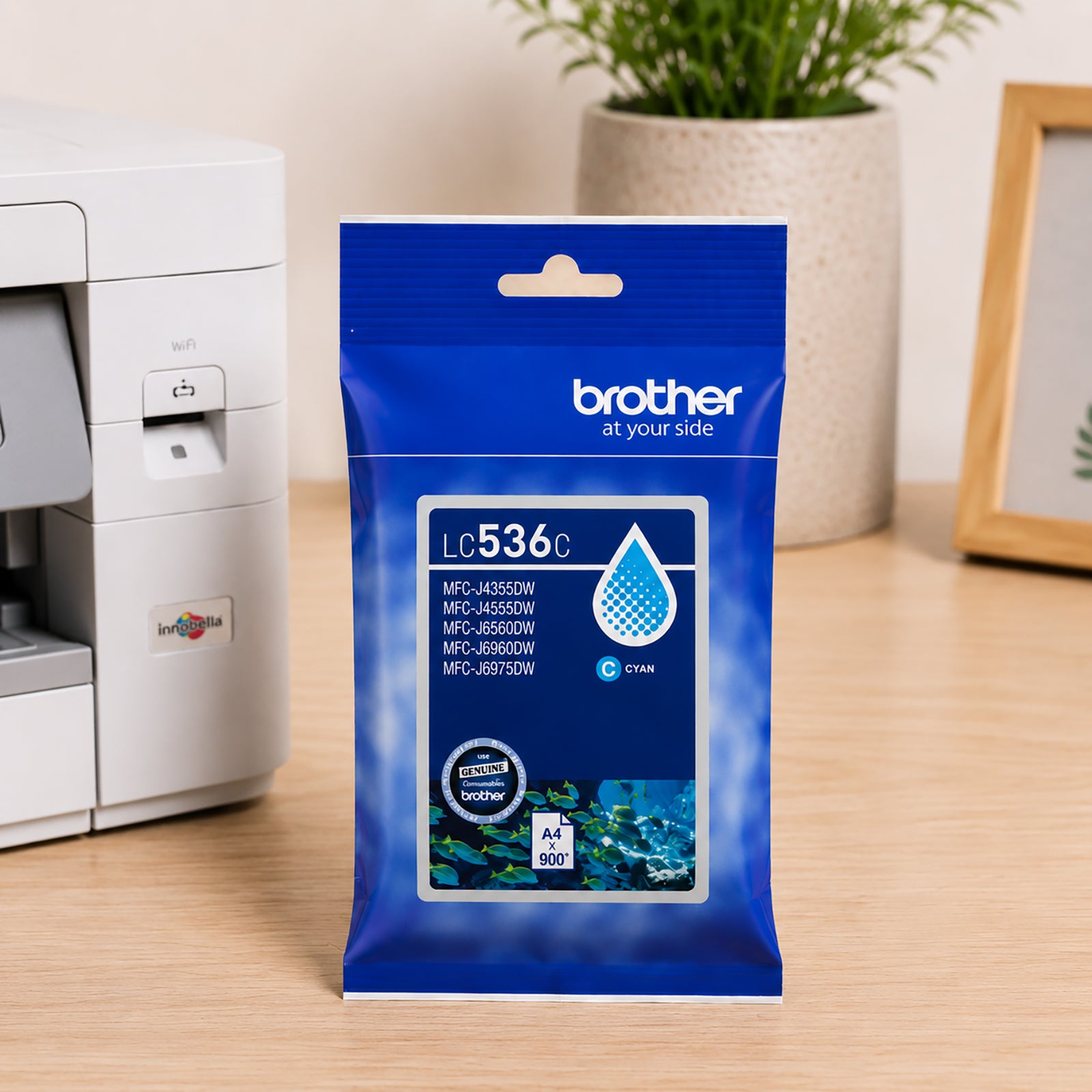 Front facing view of the Brother LC536C Cyan Ink Cartridge positioned on a clean office desk beside a printer and decorative workspace accessories. The packaging clearly displays printer compatibility and page yield information, reinforcing dependable genuine Brother performance.