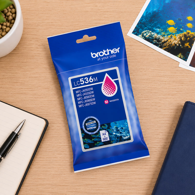 Top down desktop scene featuring the Brother LC536M Magenta Ink Cartridge alongside notebooks, office accessories and colourful printed photographs. The image showcases the cartridge's suitability for creative projects, photo printing and high quality colour document production.