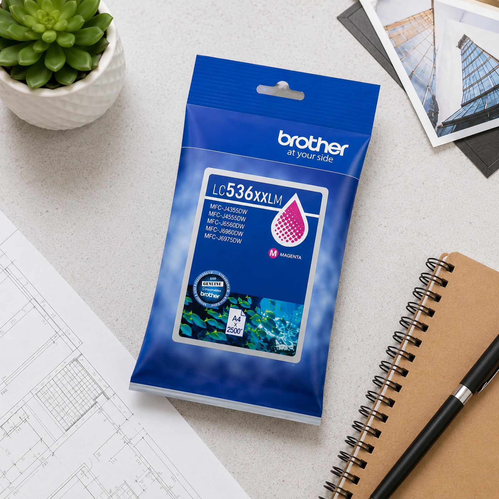 Top view of the Brother LC536XXLM super high yield magenta ink cartridge retail packaging placed on a desk beside architectural plans, a notebook and office accessories. The image showcases genuine Brother packaging and suitability for professional workplace printing environments.