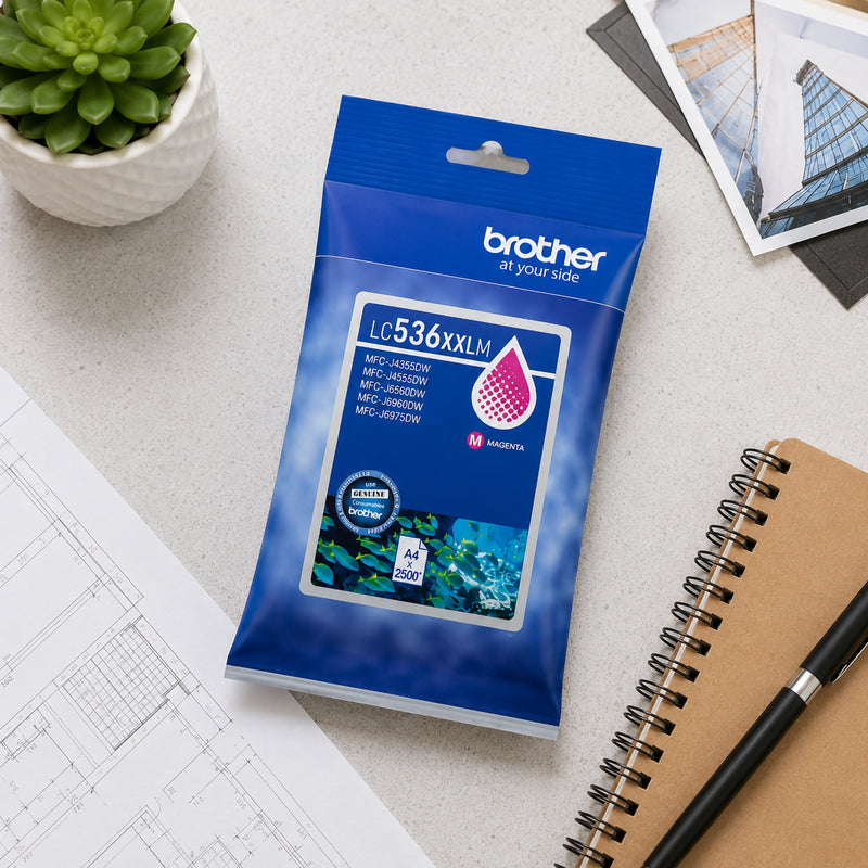 Top view of the Brother LC536XXLM super high yield magenta ink cartridge retail packaging placed on a desk beside architectural plans, a notebook and office accessories. The image showcases genuine Brother packaging and suitability for professional workplace printing environments.