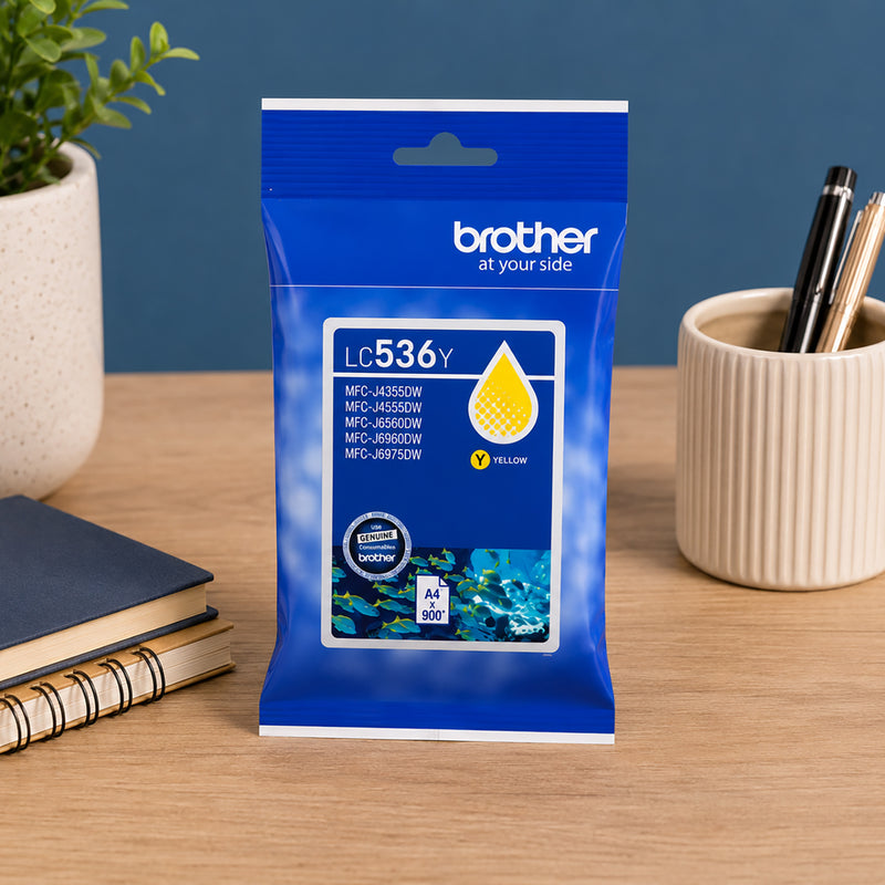 Front facing product presentation of the Brother LC536Y Yellow Ink Cartridge positioned on a clean office desk with books and stationery. The packaging clearly displays printer compatibility details and emphasises dependable genuine Brother ink performance for consistent colour printing.