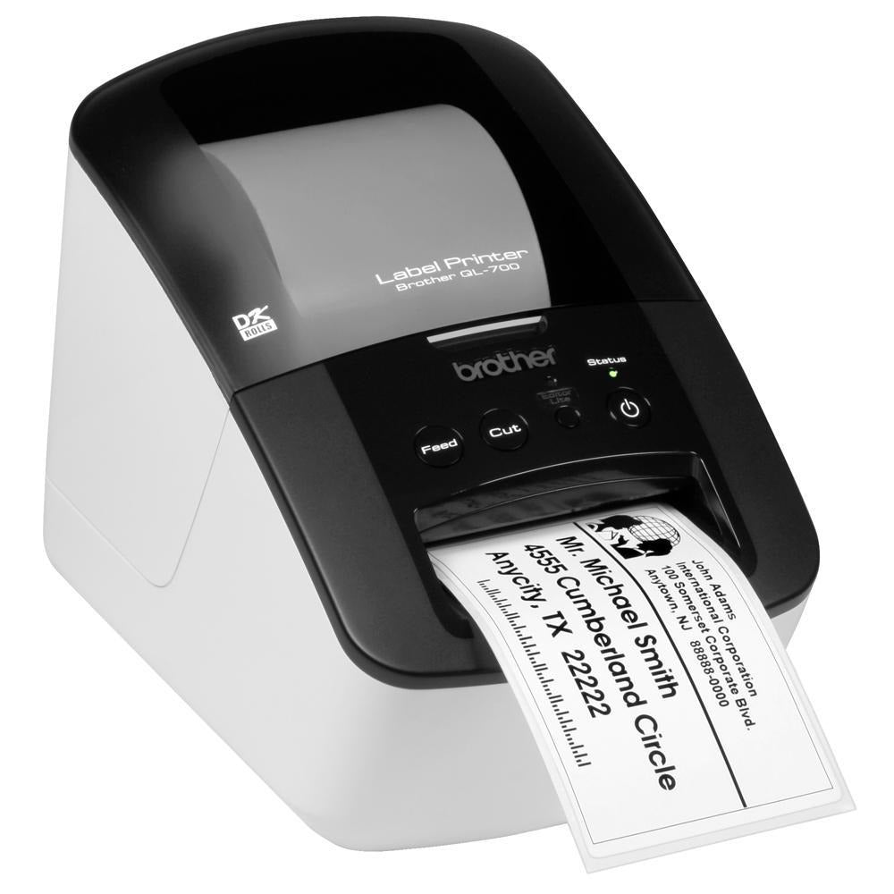 Brother QL-700 label printer viewed from the front left on a white background, showing the brother wordmark, the green status light and a printed white address label with sender and recipient details emerging from the front slot.