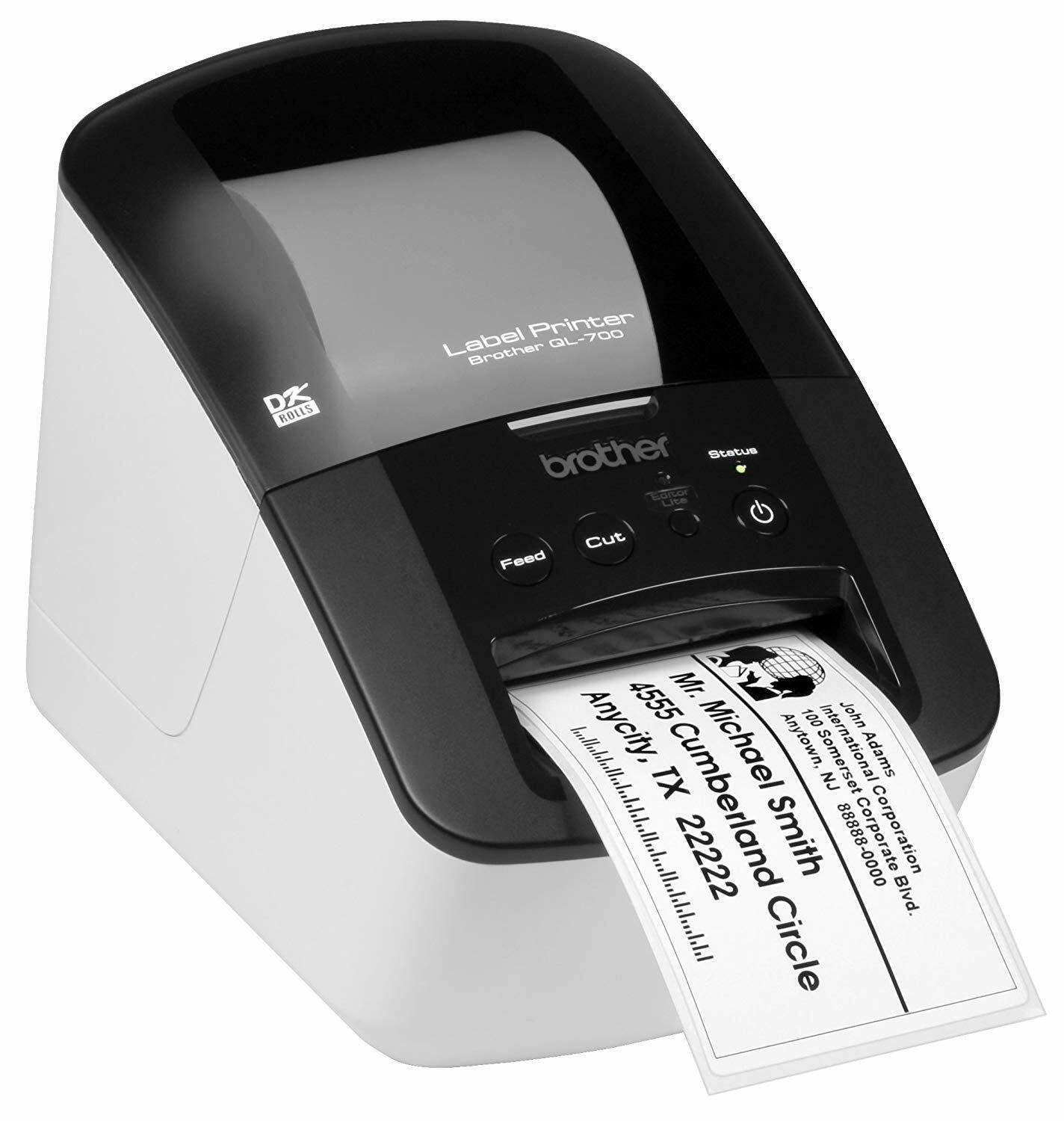 Brother QL-700 label printer angled to the right on a white background, white body with a black top and smoked grey roll cover reading Label Printer Brother QL-700, and a printed white address label feeding out of the front slot.
