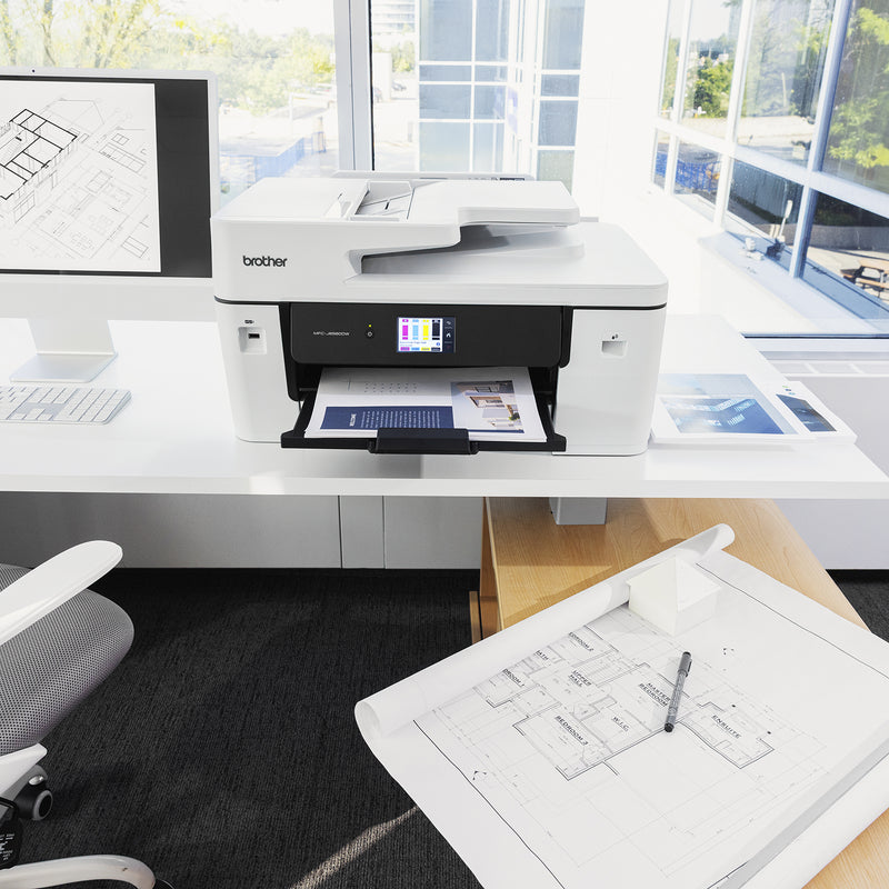 Front facing view of the Brother MFC J6560DW operating in a bright office workspace. A colour document is being printed while technical drawings and design plans are displayed nearby, showcasing the printer's ability to support creative, engineering and business applications.