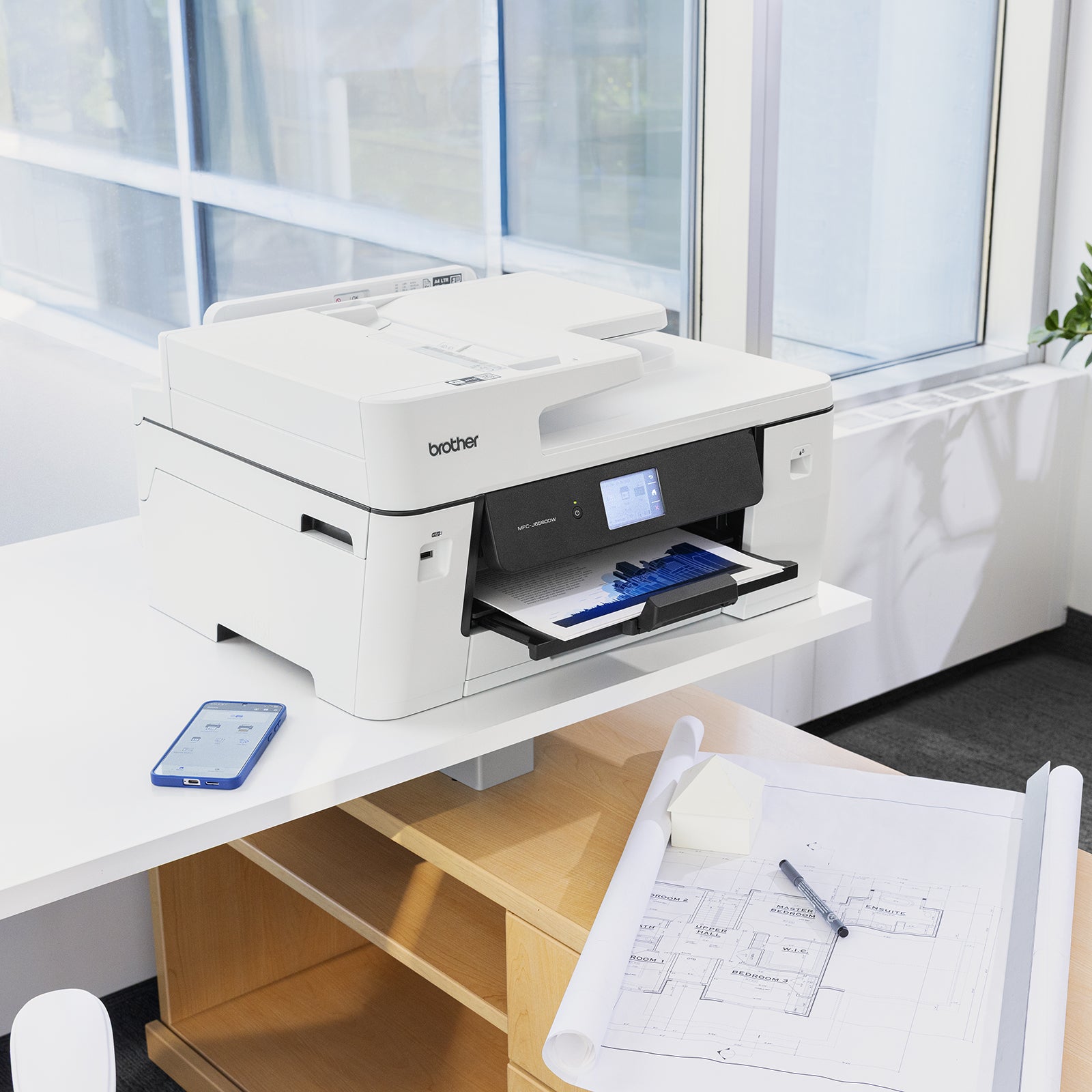 Brother MFC J6560DW A3 multifunction inkjet printer positioned in a modern office environment printing a full colour business document. The printer sits on a white workstation beside architectural plans and a mobile phone, highlighting its suitability for professional offices requiring A3 printing, scanning, copying and wireless productivity.