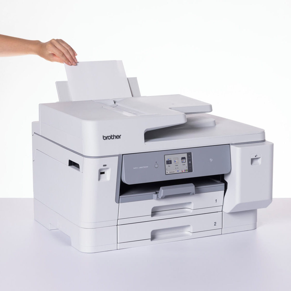 Hand loading a single sheet into the rear multi purpose tray of the Brother MFC-J6975DW, with the front touchscreen and paper trays 1 and 2 visible.