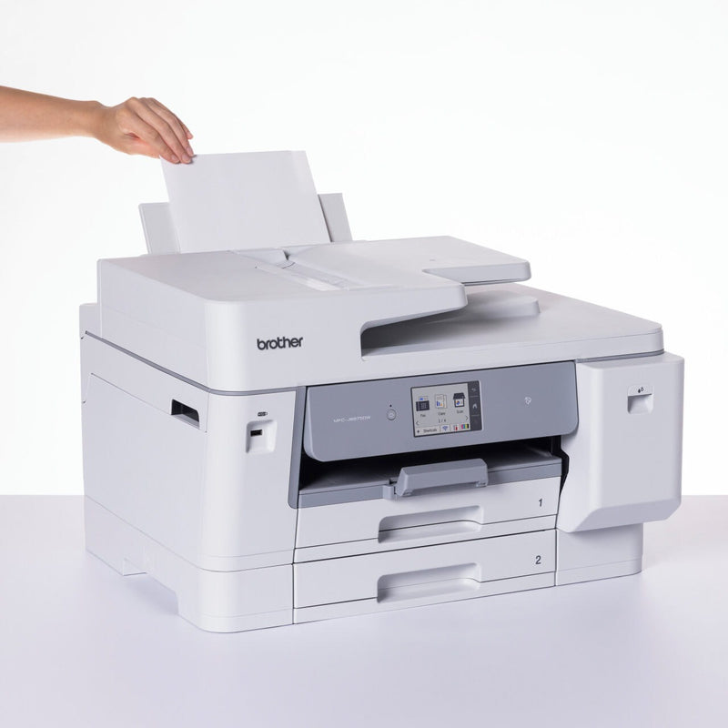 Hand loading a single sheet into the rear multi purpose tray of the Brother MFC-J6975DW, with the front touchscreen and paper trays 1 and 2 visible.