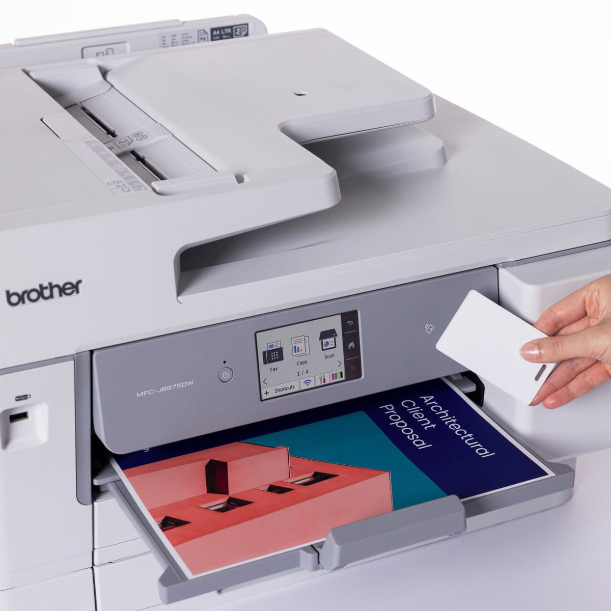 Hand holding a white card to the NFC reader on the front of the Brother MFC-J6975DW, with a colour Architectural Client Proposal page in the output tray below the touchscreen.