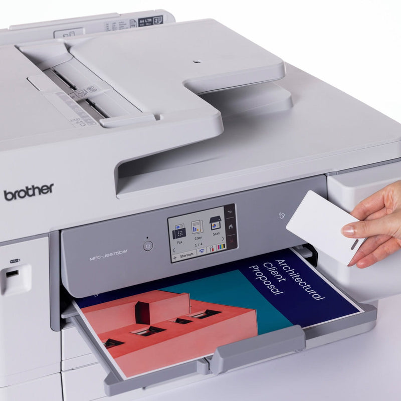 Hand holding a white card to the NFC reader on the front of the Brother MFC-J6975DW, with a colour Architectural Client Proposal page in the output tray below the touchscreen.