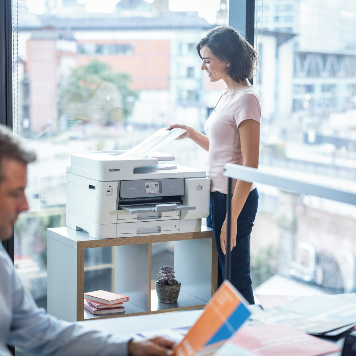 Woman in a pink top collecting a print from a Brother MFC-J6975DW on a low white shelving unit in front of a glass office wall, with a colleague reading documents at a desk.