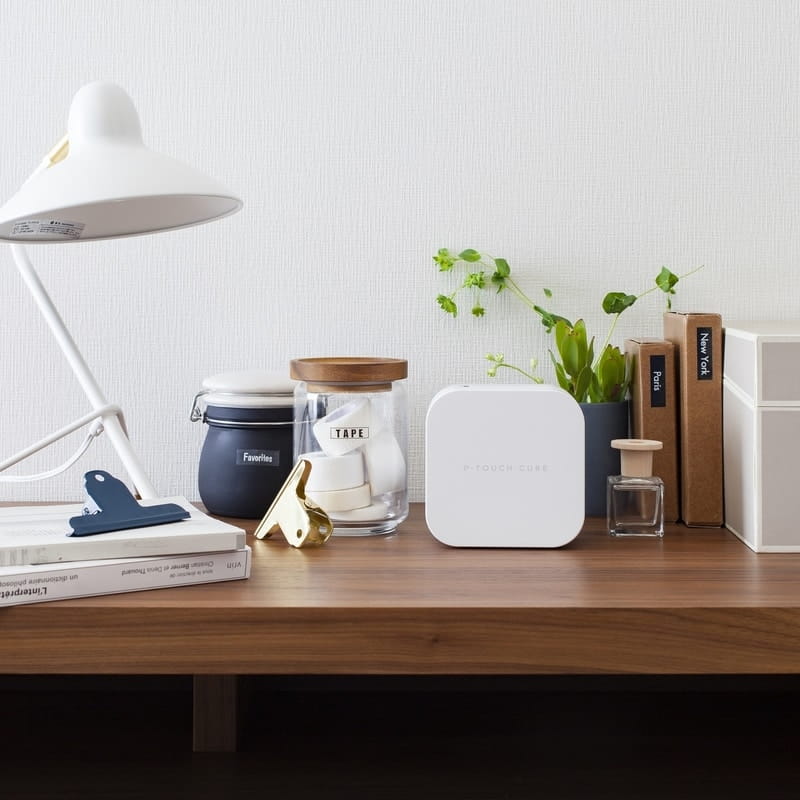 Brother PT-P300BT P-Touch Cube label maker standing upright on a walnut desk beside a white lamp, stacked books and glass jars labelled Favourites and Tape, showing the compact white cube in a styled workspace setting