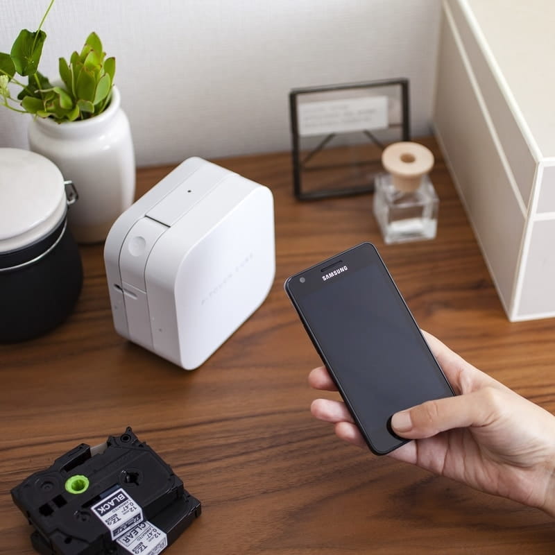 Hand holding a smartphone next to the Brother PT-P300BT P-Touch Cube on a timber desk, with two black TZe tape cassettes in the foreground, showing how a label is designed in the app and sent to the printer over Bluetooth