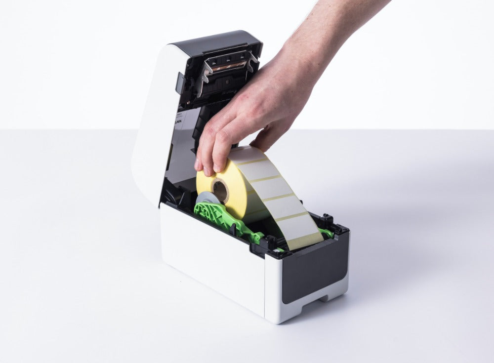 Hand lowering a roll of yellow-backed white die cut labels into the open Brother TD-2320D, with the lid raised and the green roll guides visible.