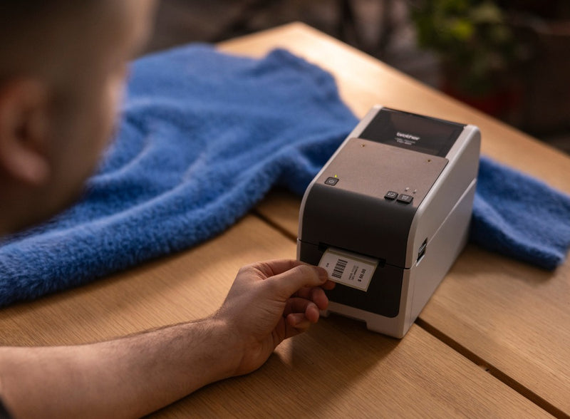 Person at a wooden retail counter tearing a printed barcode price label from the Brother TD-2320D, with a blue knitted jumper laid on the bench.