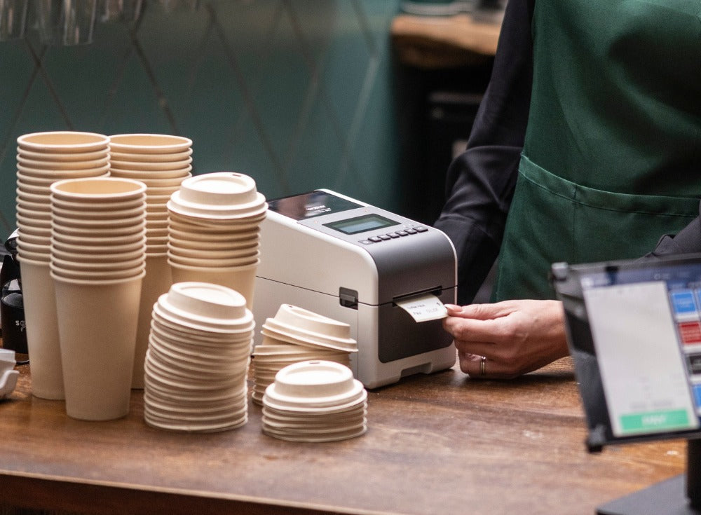 Barista in a green apron tearing a printed label from a Brother TD-2350D on a cafe counter beside stacks of takeaway cups and lids, with a tablet point of sale in the foreground.