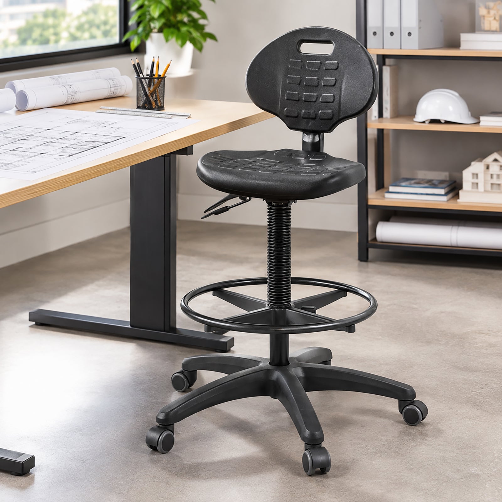 Buro Enso drafting chair positioned beside an architectural workstation with plans and drawing tools, showing the elevated seating configuration, black circular foot ring, tall gas lift, mobile base and practical moulded chair design.