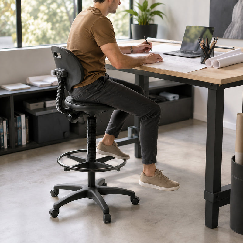 Lifestyle view of a Buro Enso Chair converted for drafting use with the black architectural base kit, showing a seated user working at an elevated design desk with the circular foot ring and extended gas lift clearly visible.