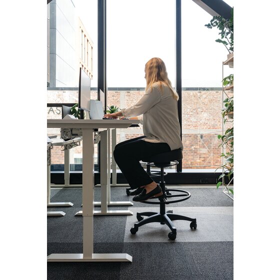 Person seated on a black Buro Polo drafting stool at a raised white standing desk in an office beside tall windows