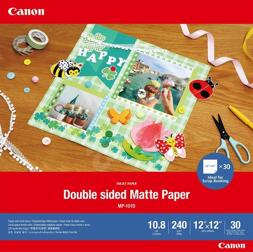 Canon Double Sided Matte Paper MP-101D craft paper pack shown on a wooden desk with scrapbooking supplies. The red and blue packaging prominently displays the Canon brand and product details including 10.