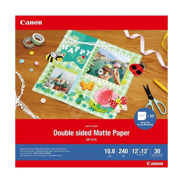 Canon Double Sided Matte Paper MP-101D craft paper pack shown on a wooden desk with scrapbooking supplies. The red and blue packaging prominently displays the Canon brand and product details including 10.