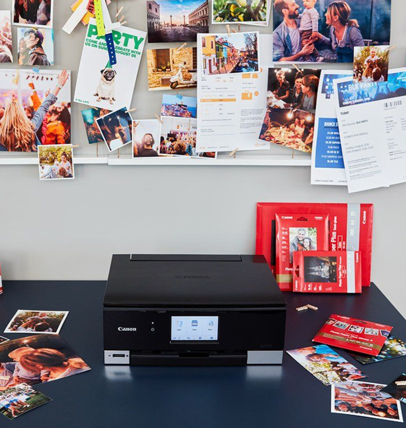 A Canon all-in-one printer sits on a dark desk in an office-like setting, surrounded by printed photos and red Canon-branded boxes against a gray wall with pinned papers.