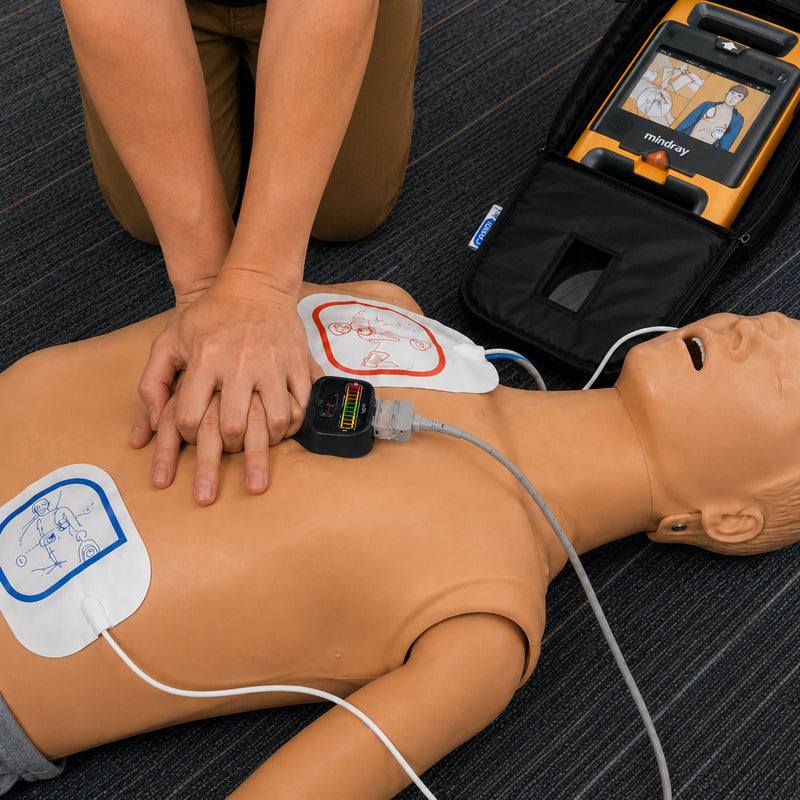 CardiAct Mindray Soft Carry Case opened beside a CPR training manikin during a simulated cardiac emergency, with a Mindray AED operating inside the case while a rescuer performs chest compressions. Electrode pads and connected leads demonstrate how the carry case supports rapid access to the defibrillator during emergency response training.