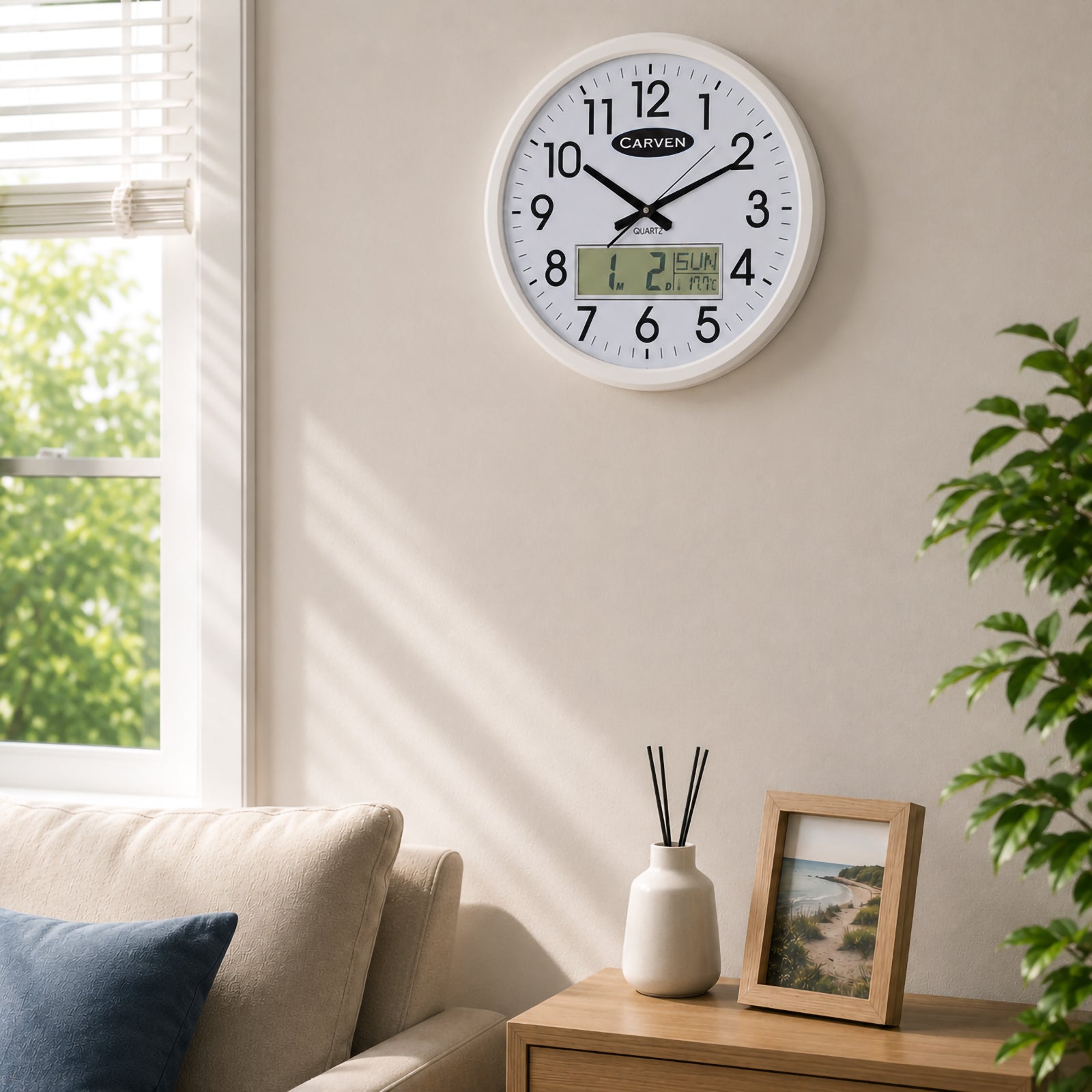 The Carven White Frame LCD Calendar Wall Clock is mounted above a cabinet beside decorative plants and home accessories in a contemporary lounge room. Natural sunlight highlights the large analogue display and digital calendar, demonstrating its suitability for modern home and office interiors.