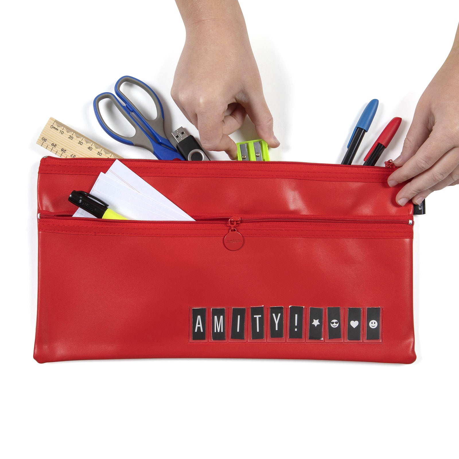 Hands organise school supplies into the Celco Large 2 Zip Name Pencil Case against a white background. Scissors, rulers, pens, highlighters, paper and sharpeners demonstrate the generous storage capacity and dual zip compartments for organised stationery.