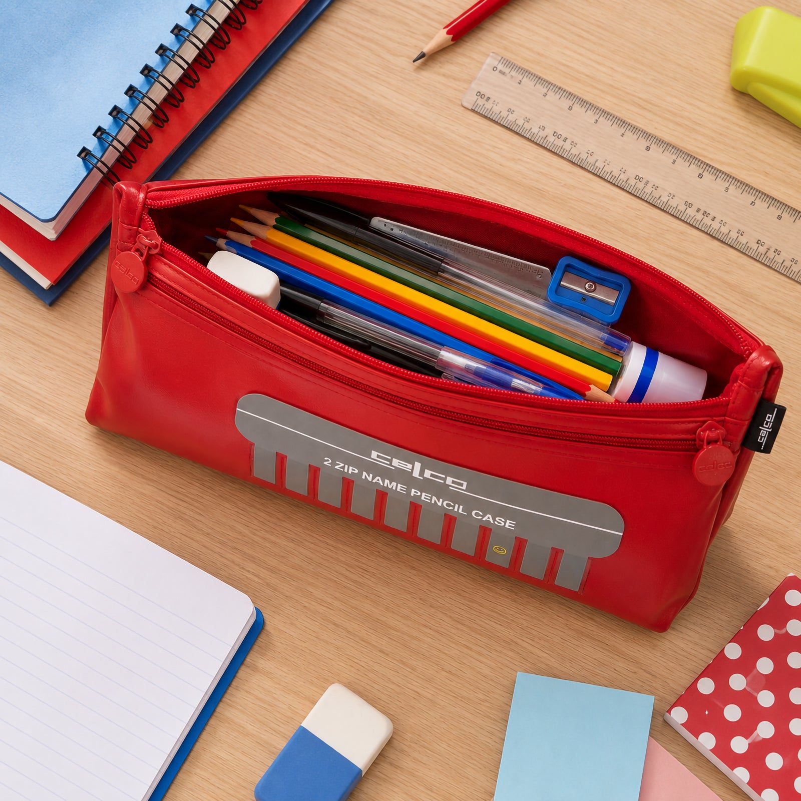 Top view of the Celco Large 2 Zip Name Pencil Case opened to reveal coloured pencils, pens, glue stick, sharpener, ruler and other stationery neatly organised inside. School notebooks, sticky notes and classroom supplies surround the pencil case on a wooden desk.