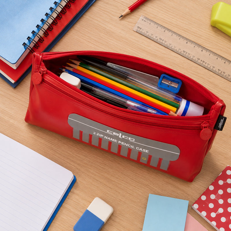 Top view of the Celco Large 2 Zip Name Pencil Case opened to reveal coloured pencils, pens, glue stick, sharpener, ruler and other stationery neatly organised inside. School notebooks, sticky notes and classroom supplies surround the pencil case on a wooden desk.