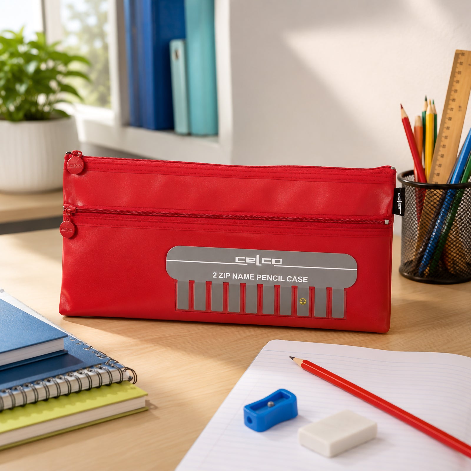 The Celco Large 2 Zip Name Pencil Case stands upright on a study desk beside notebooks, pencils, an eraser and sharpener. The vibrant red finish and personalised name window are highlighted in a bright learning environment suitable for students and home study.