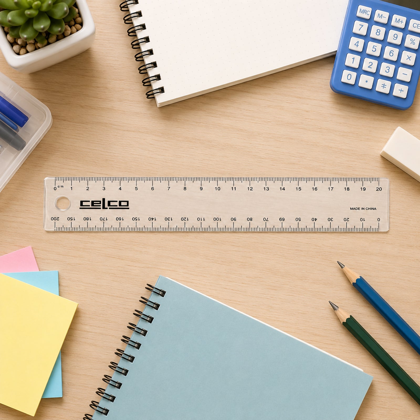 Top view of the Celco 20cm clear ruler displayed neatly on a wooden desk surrounded by notebooks, pencils, sticky notes, a calculator and stationery accessories. The transparent ruler is positioned prominently, highlighting its clear metric graduations.