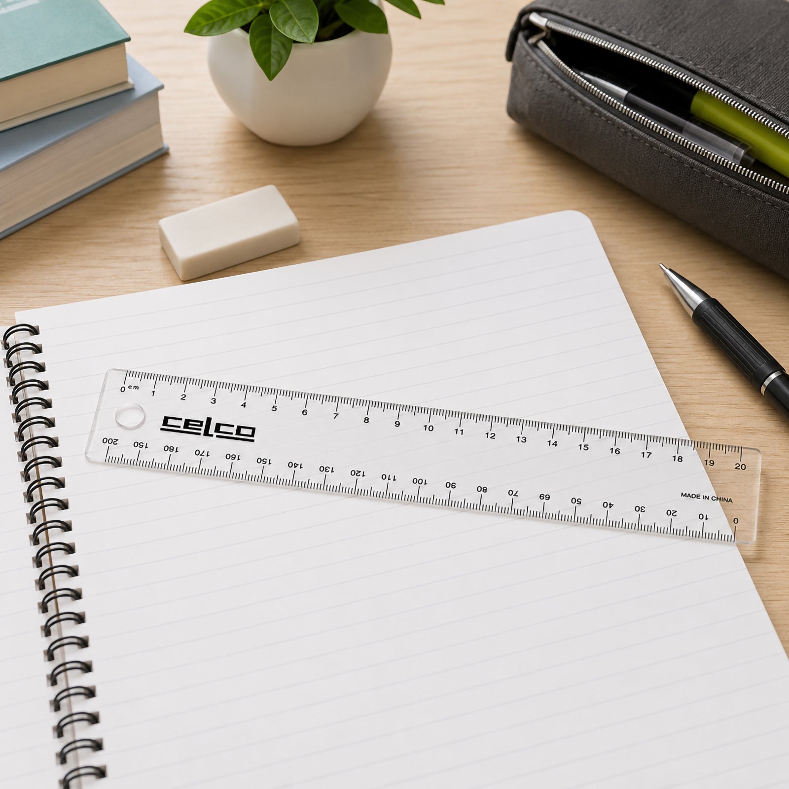 The Celco 20cm clear ruler rests diagonally across an open lined notebook on a tidy office desk. Nearby books, a pen, pencil case, plant and eraser create an organised workspace that showcases the ruler as an everyday measuring tool.