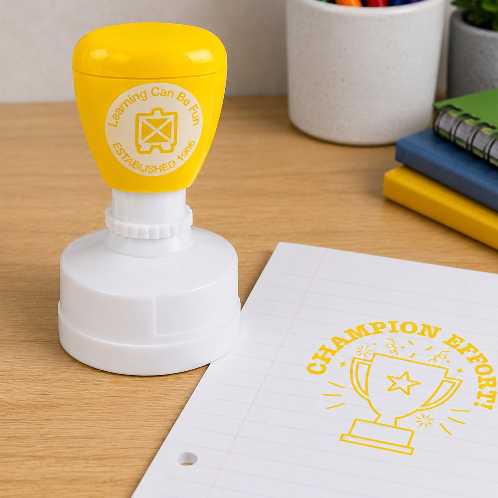Learning Can Be Fun Champion Effort self inking merit stamp displayed beside lined paper featuring a bright yellow trophy award impression. The stamp celebrates determination, persistence and positive student achievement in the classroom.