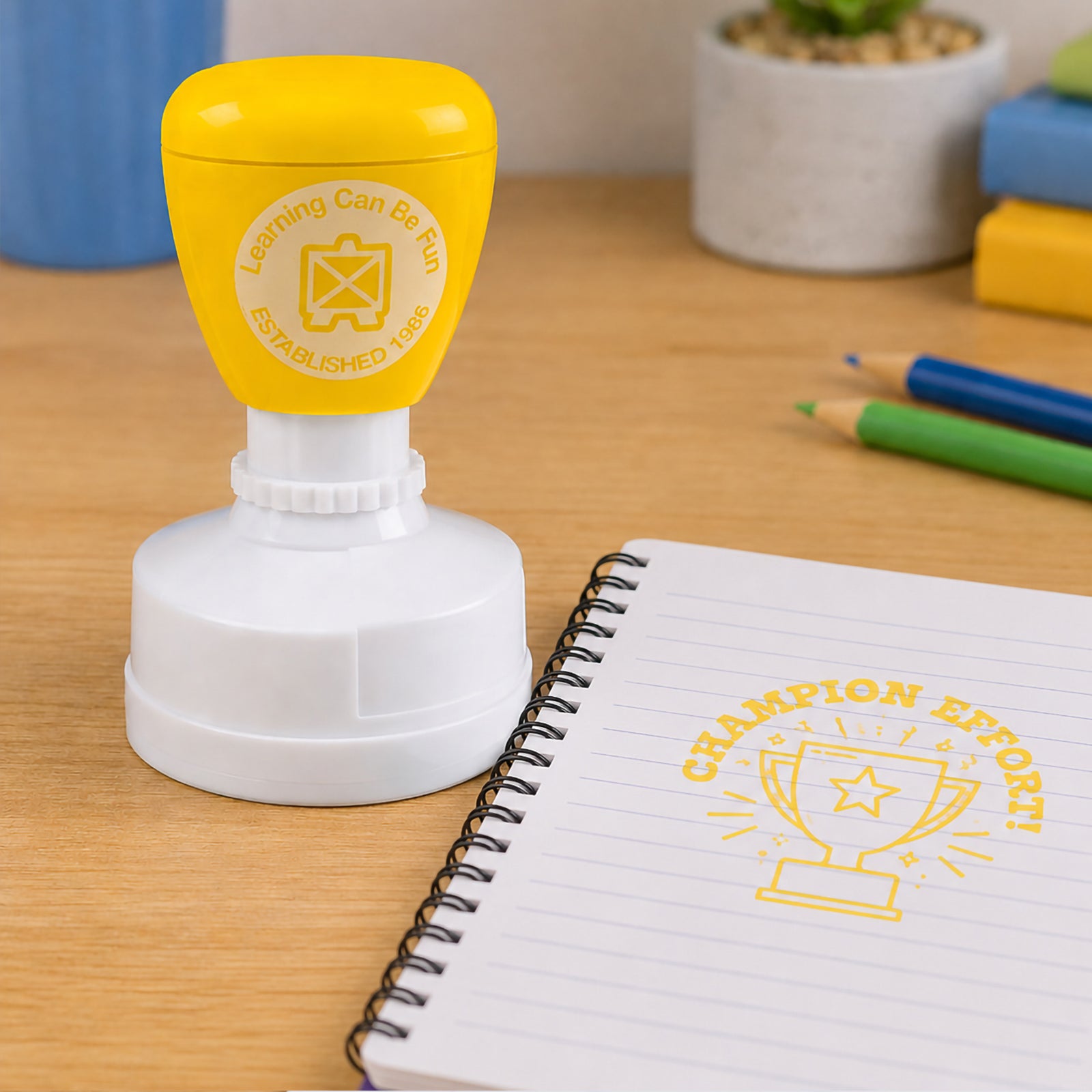 Champion Effort merit stamp positioned next to a spiral notebook and coloured pencils on a classroom desk. The bold yellow reward design provides encouraging recognition for students who consistently try their best.
