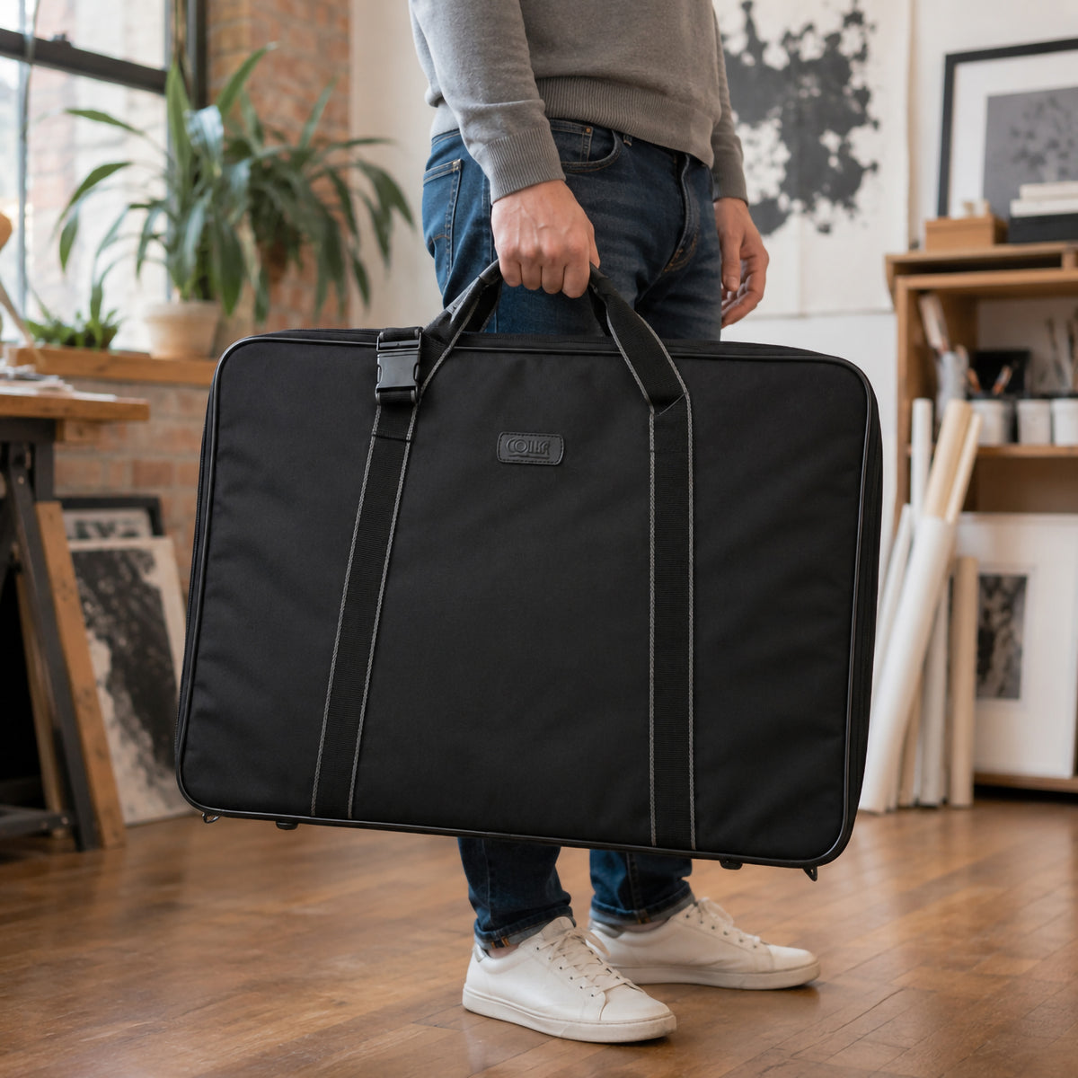 Colby Art A2 soft sided art bag in black being carried by its handles in a creative studio environment. The large portfolio bag features a structured rectangular shape, reinforced edges, carry straps and secure closure, demonstrating a practical way for artists, students and designers to transport A2 artwork and documents.