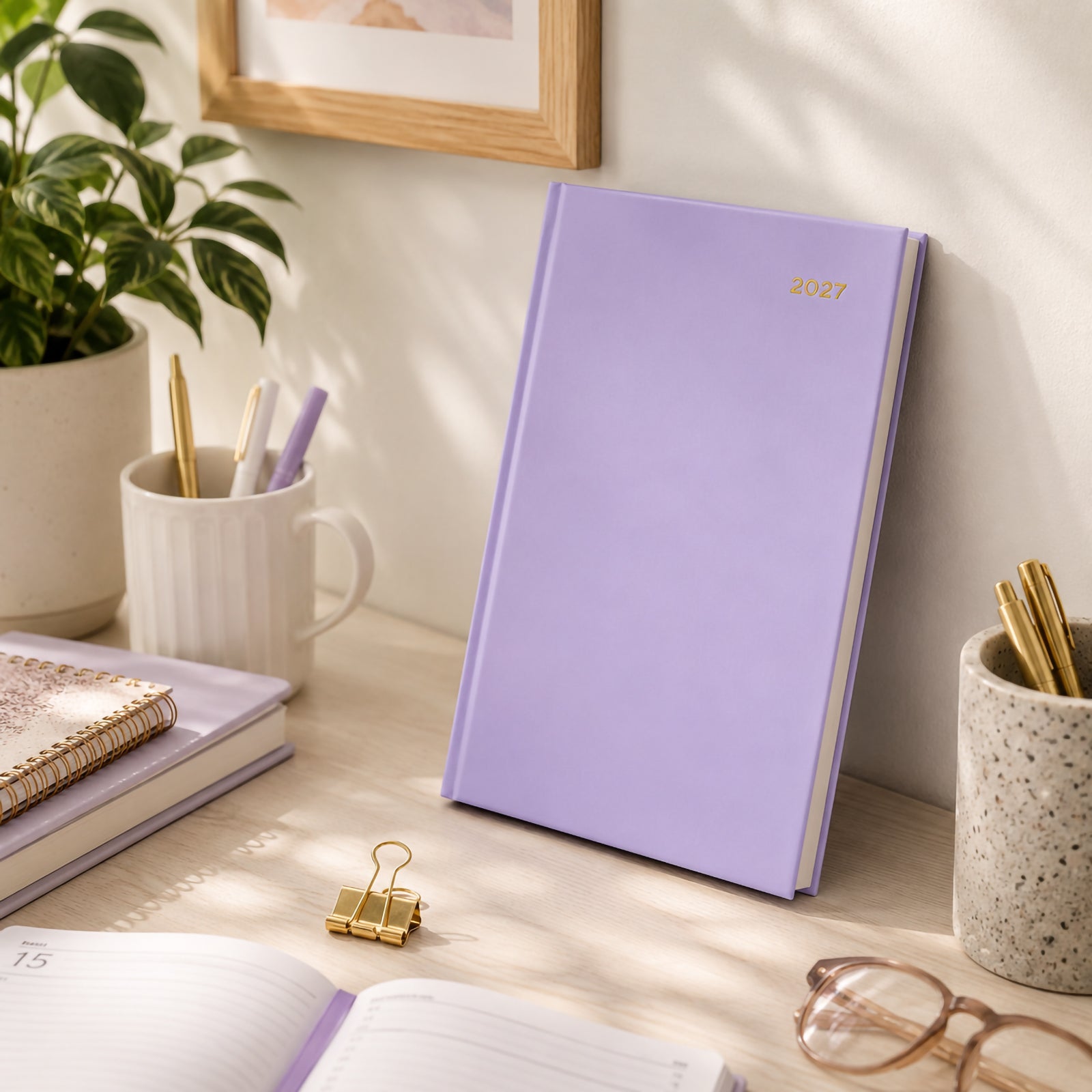 The Collins Belmont Colours 2027 A5 Day to a Page Lilac Diary stands upright on a tidy desk beside matching stationery, decorative plants and premium office accessories. Soft natural light emphasises the elegant lilac finish and contemporary workspace styling.